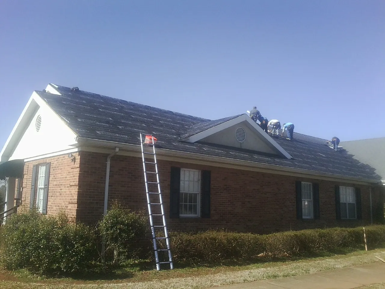 Crew tearing shingles off a brick ranch roof with a ladder set — Metal Roof Repair work by our crew in Karval