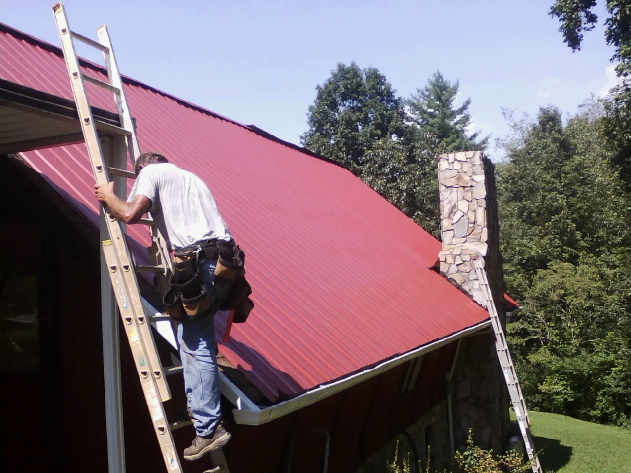 Roofer on a ladder beside a red metal roof and stone chimney — Soffit & Fascia Repair by Roofing Brothers in Karval